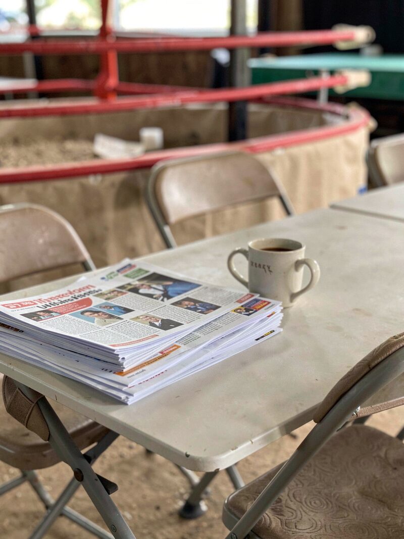 A stack of folded printed newsletters on a metal folding table beside a show catalog, a coffee cup ring on the top page, ring ropes in the background.
